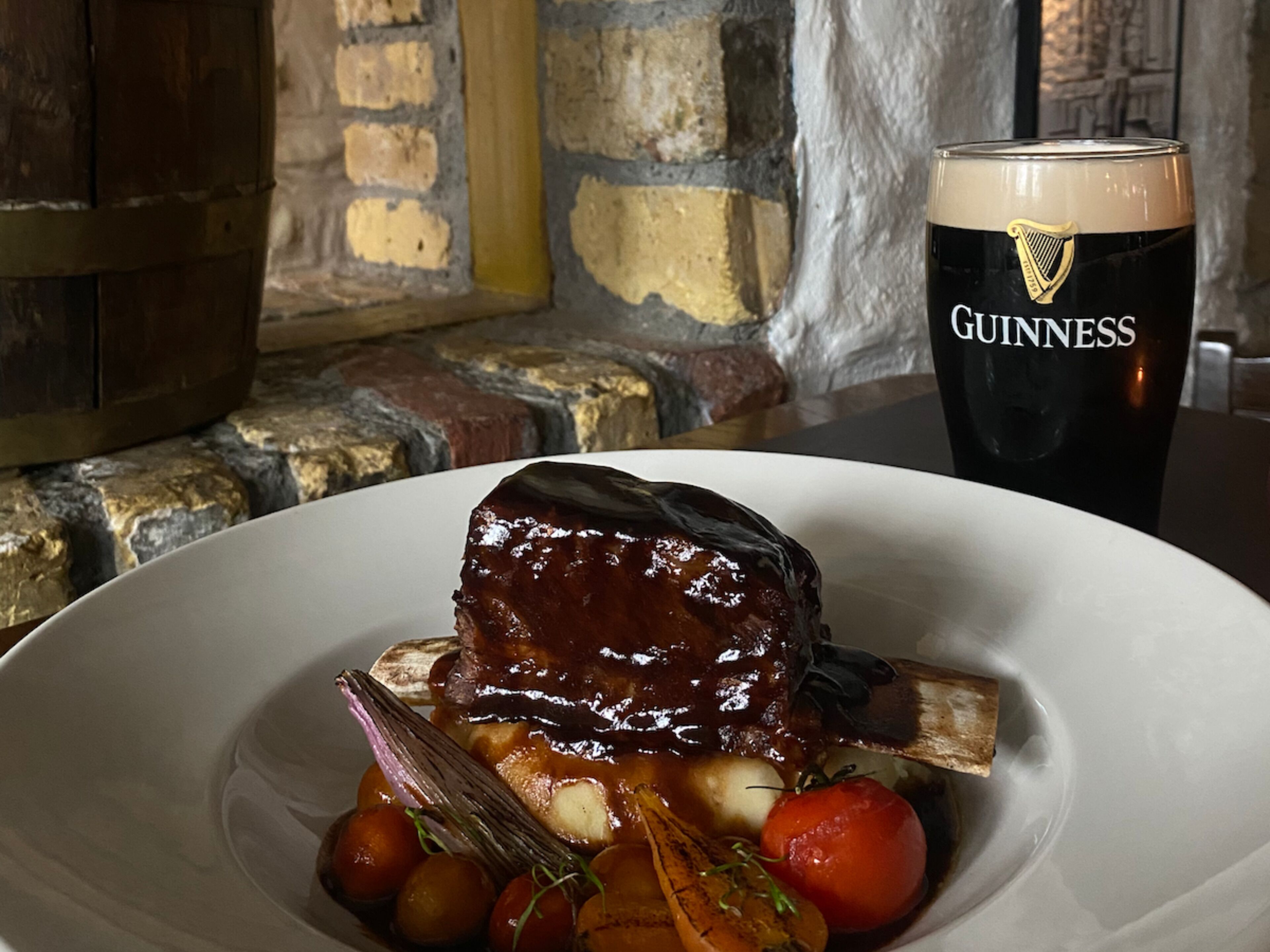 Een bord eten met een donkere geglazuurde vleesbereiding, omringd door groenten, en een glas Guinness bier.
