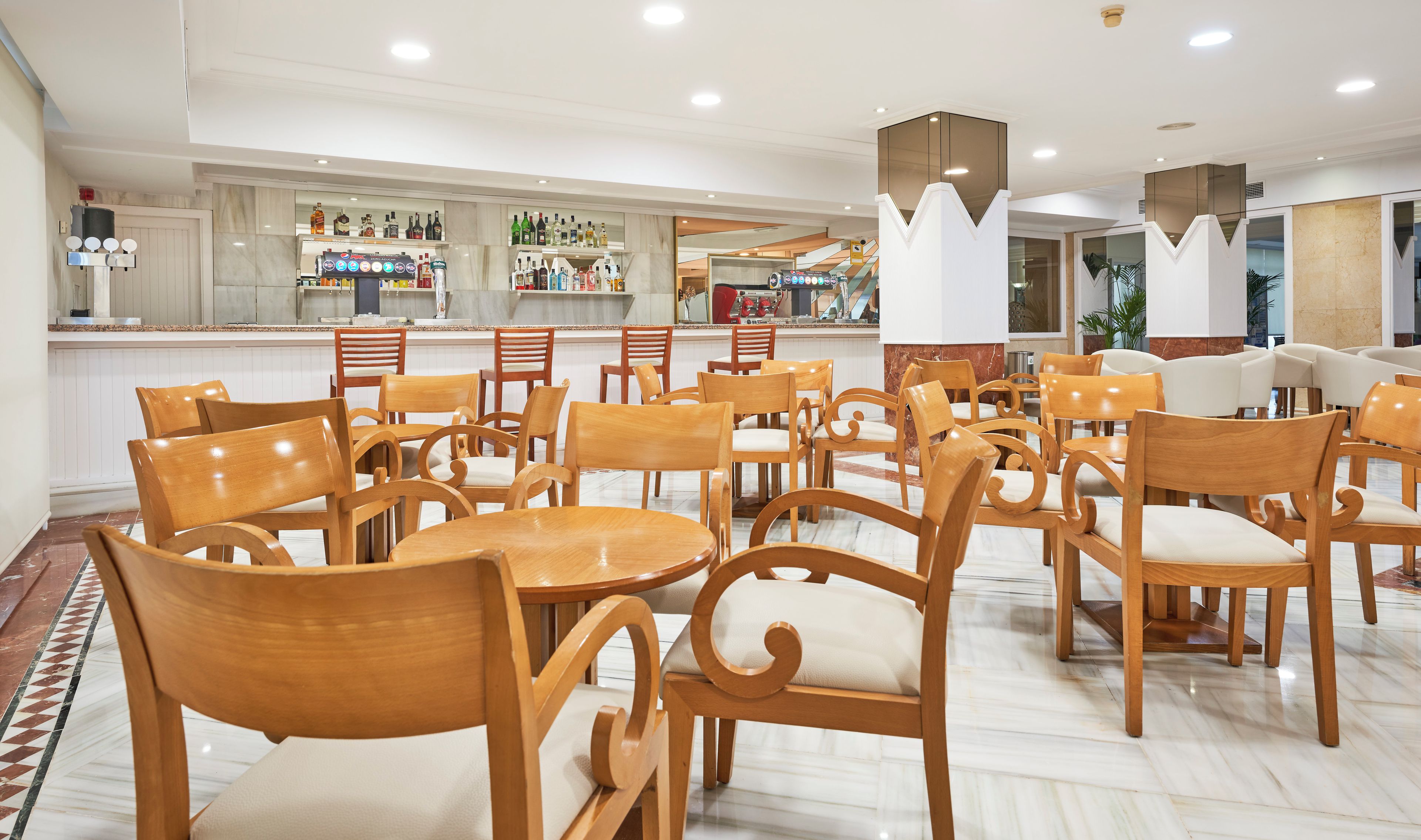 Un hall de hotel con sillas y mesas de madera, una zona de bar con diferentes botellas y un diseño de techo moderno.