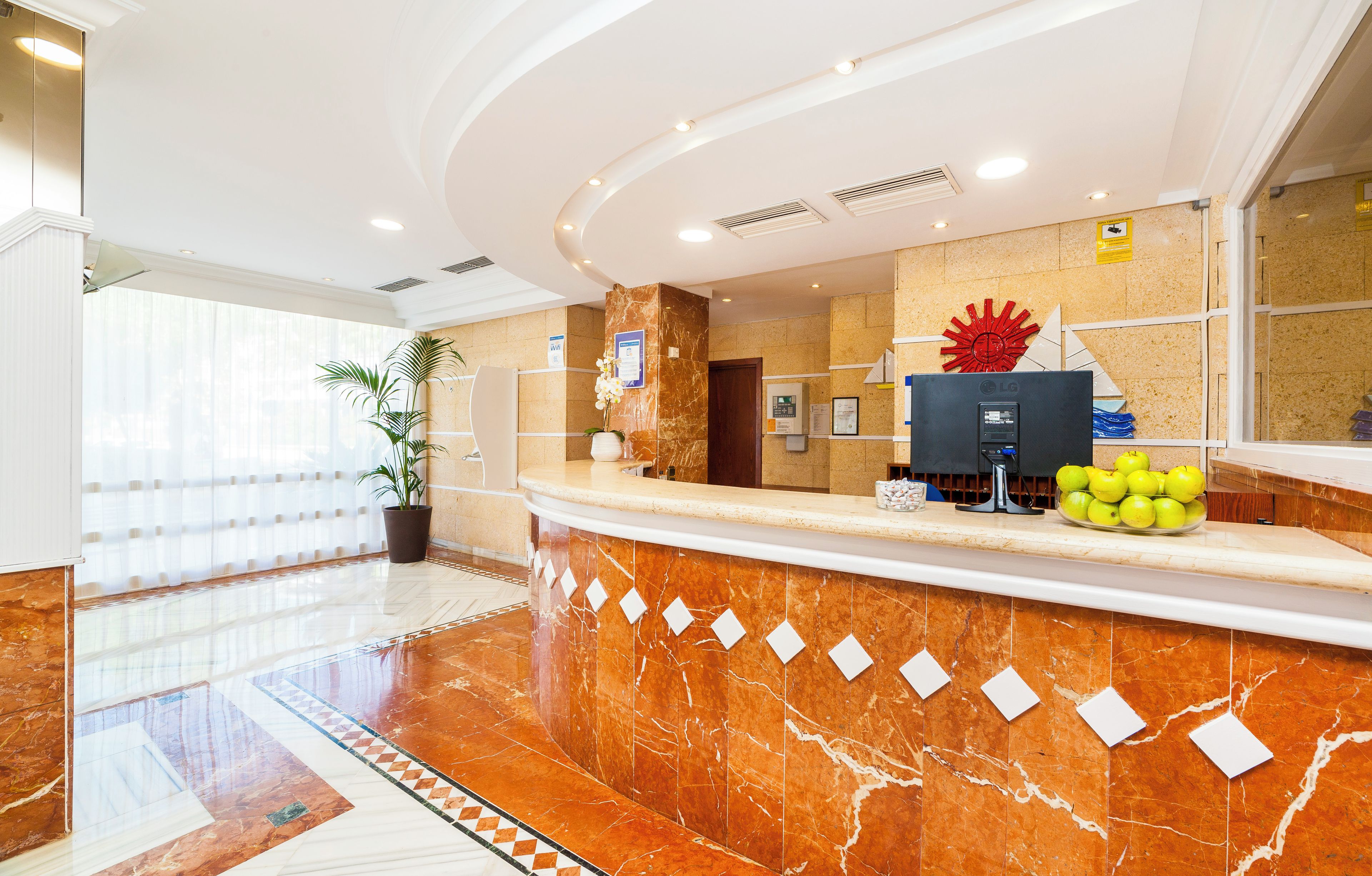 Zona de recepción de hotel con mostrador de mármol, una gran pieza decorativa roja y un frutero con manzanas.
