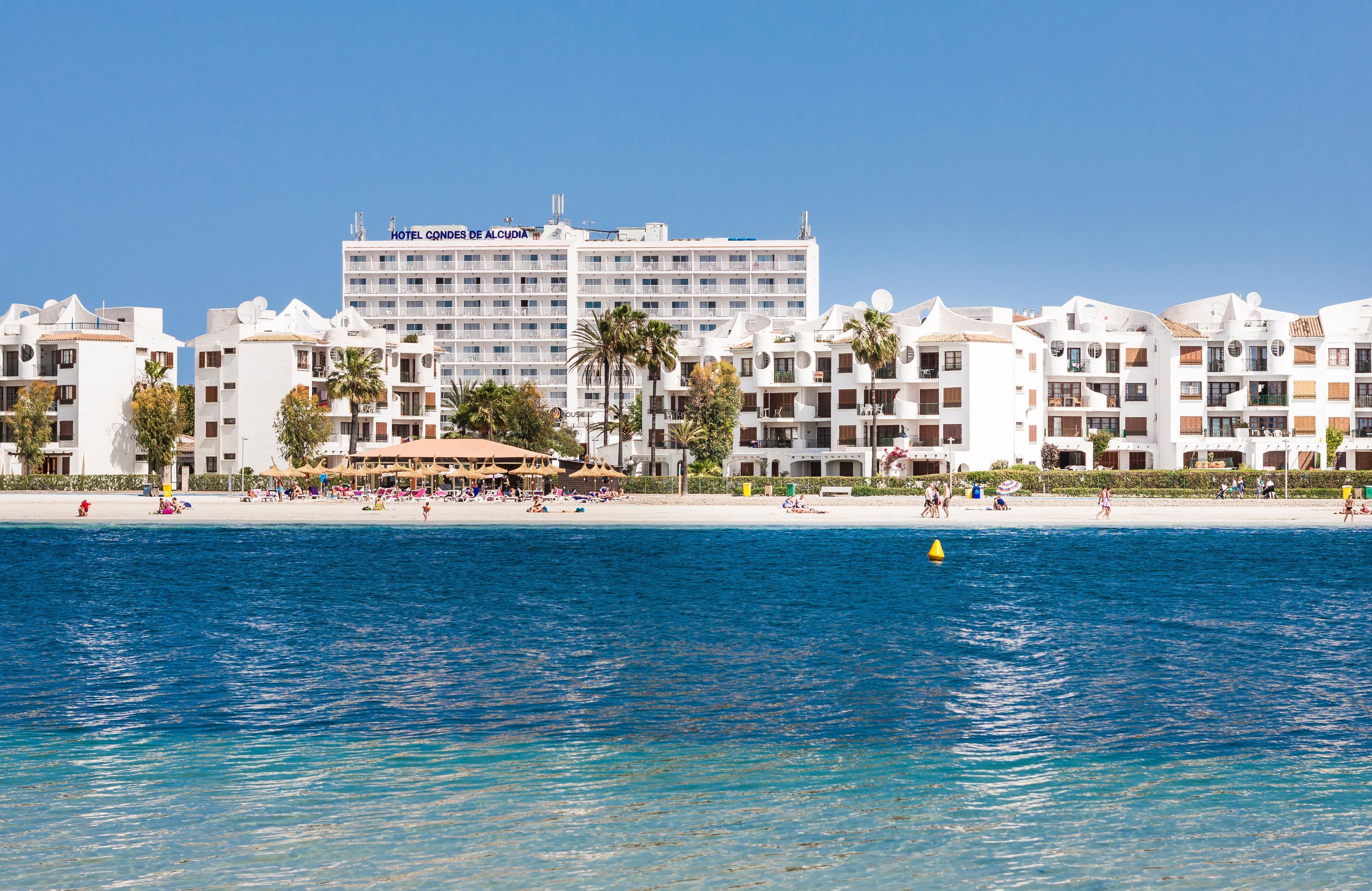 Un hotel frente a la playa con varios balcones, una playa de arena y aguas azul turquesa.