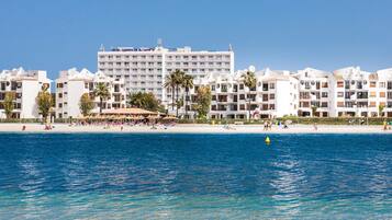 Un hotel frente a la playa con varios balcones, una playa de arena y aguas azul turquesa.