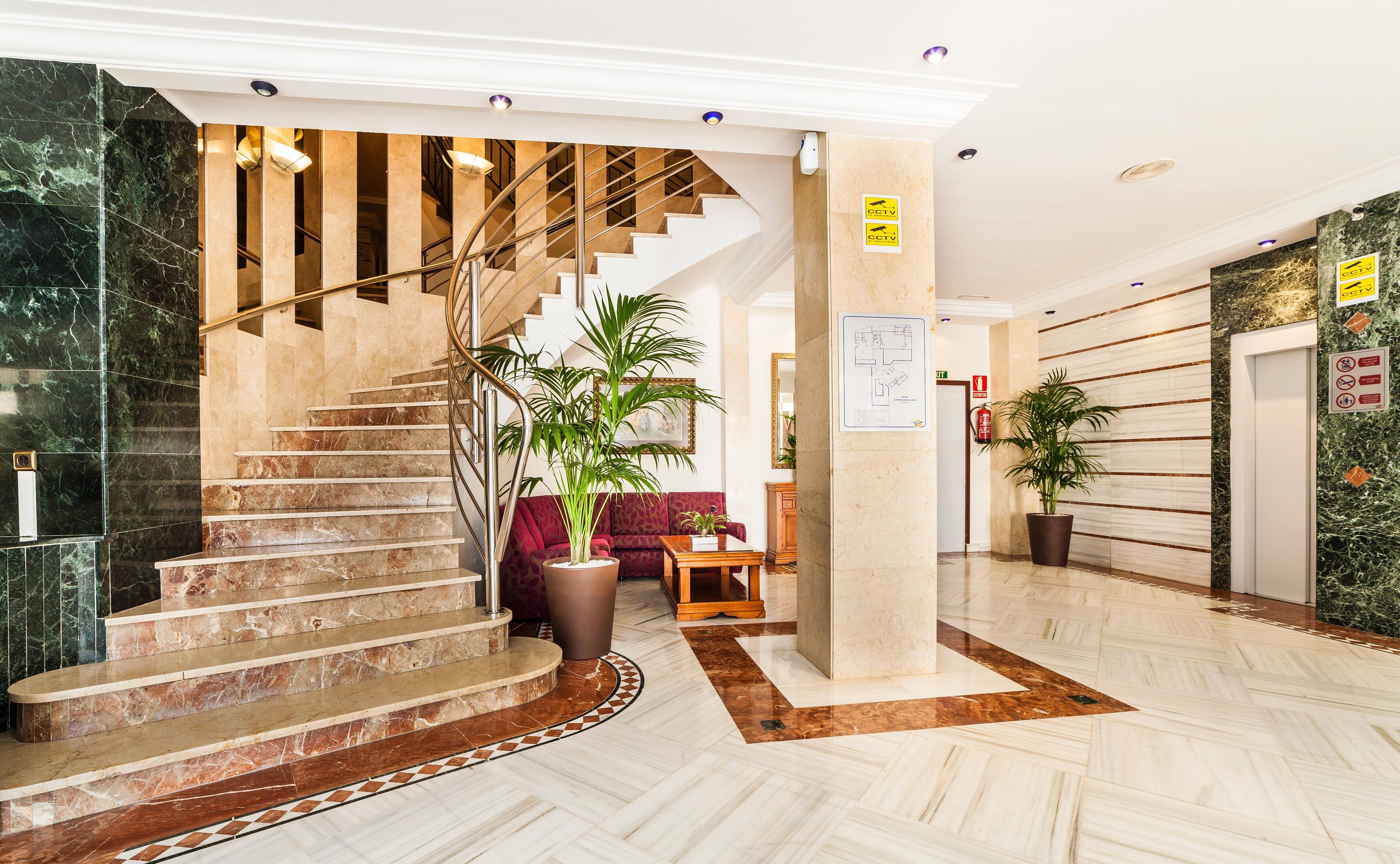 Un hall de hotel con una escalinata de mármol, un sofá rojo, una mesa de centro de madera y plantas en macetas.