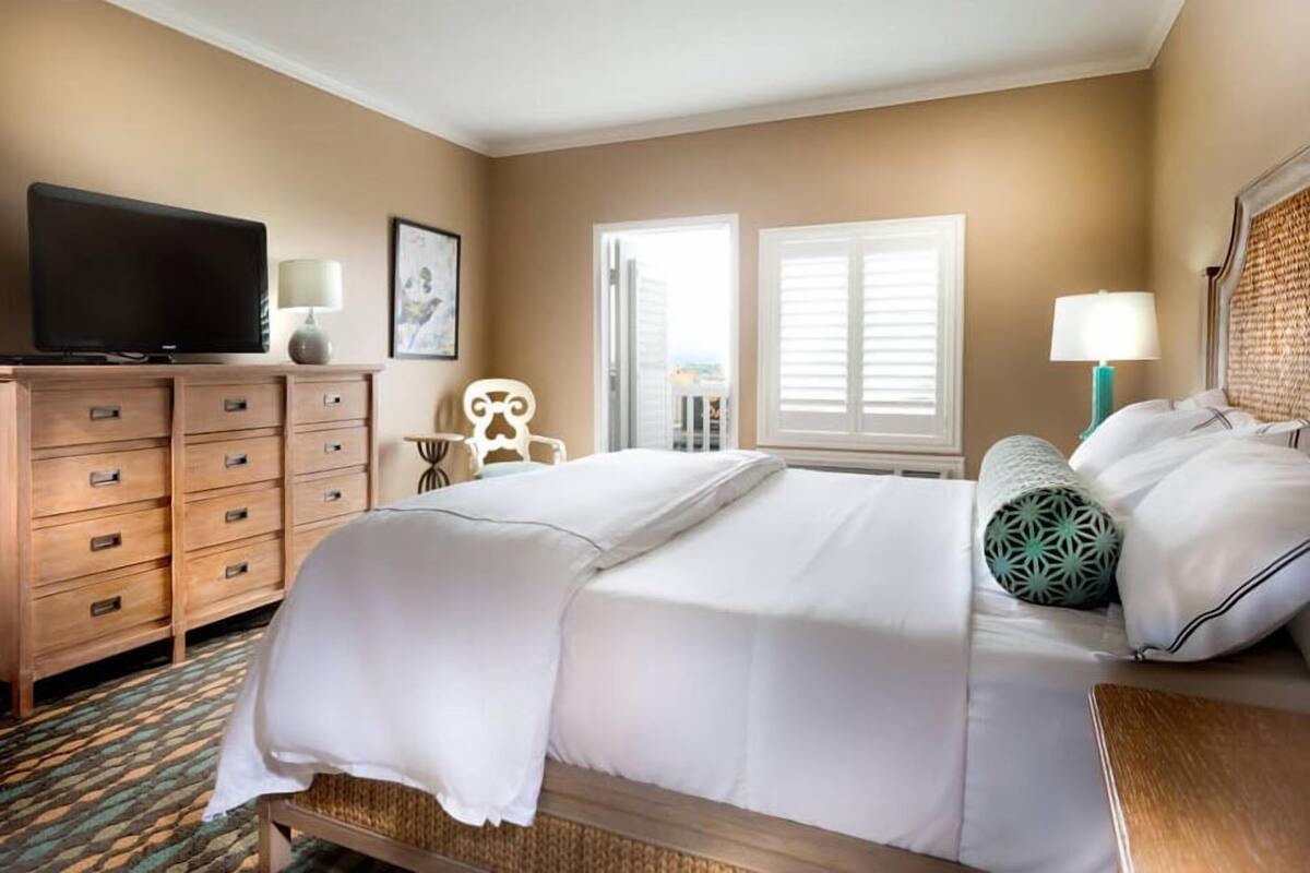 A hotel room with a bed, a television on a wooden dresser, a chair, and two windows with shutters.