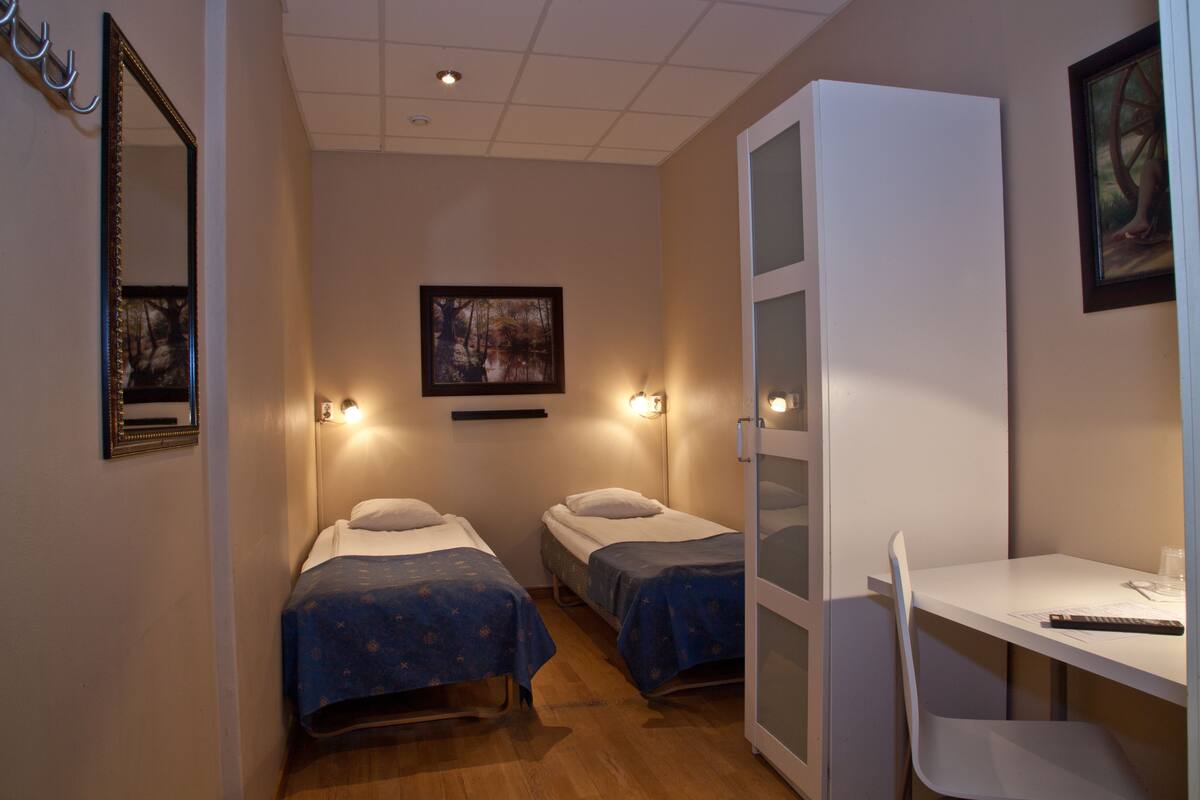 A room with two beds, a desk, and a chair. There is a painting on the wall and a mirror above the bed.