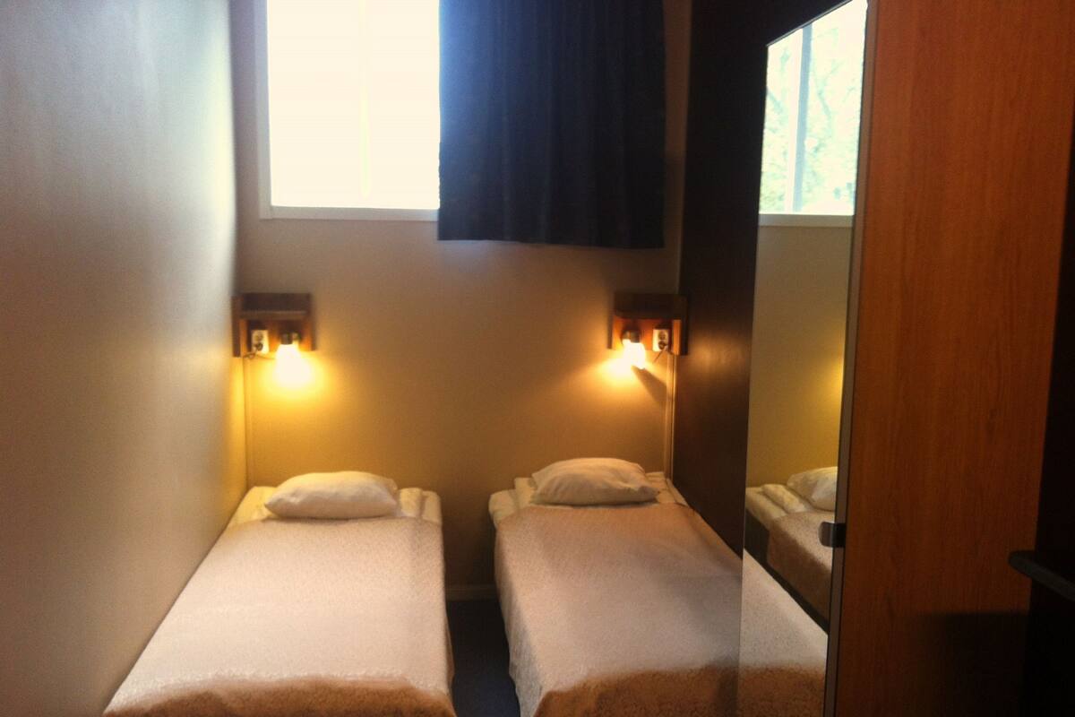 A hotel room with two single beds, each with a pillow and a nightstand. There is a window with blinds, a mirror on the wall, and a door to the right.