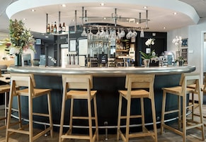 Una zona de bar con un mostrador curvo, taburetes de madera, copas de vino colgantes y una variedad de botellas y adornos.