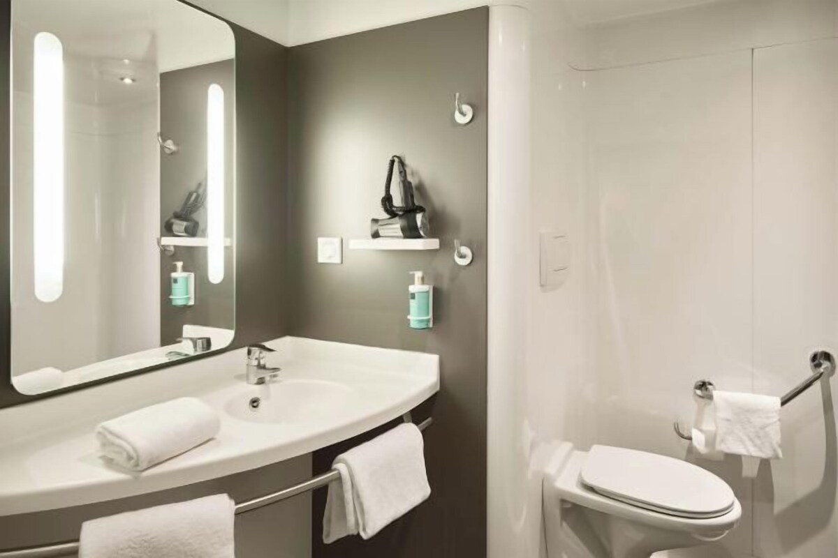 A modern bathroom with a large mirror, white sink, and toilet. There are shelves with toiletries and a towel rack.