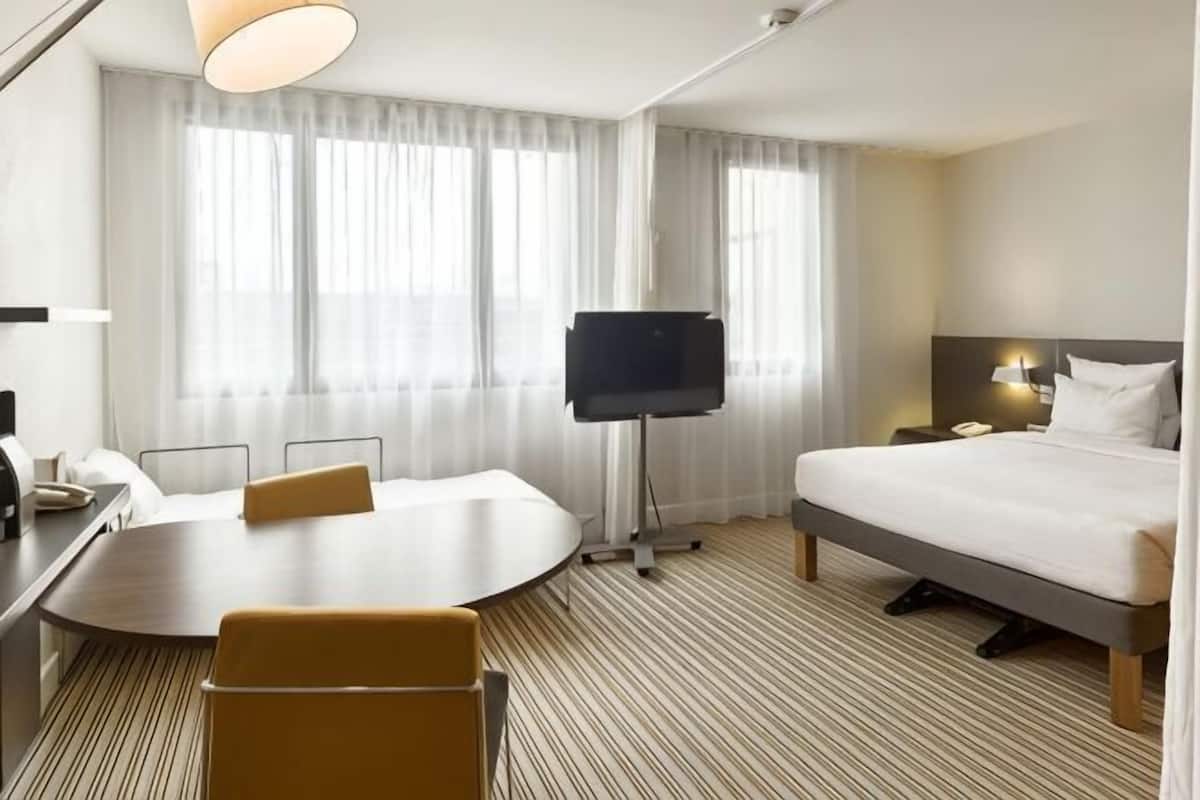 A hotel room with a bed, a desk, a chair, a TV, and a large window with curtains.