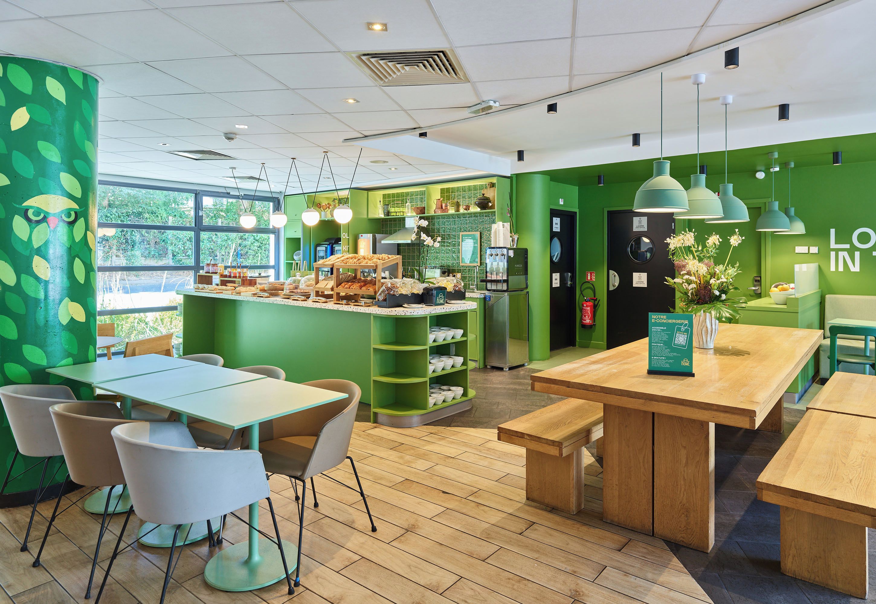 A cafe with green walls, wooden tables, and chairs. There is a counter with pastries and a coffee machine.