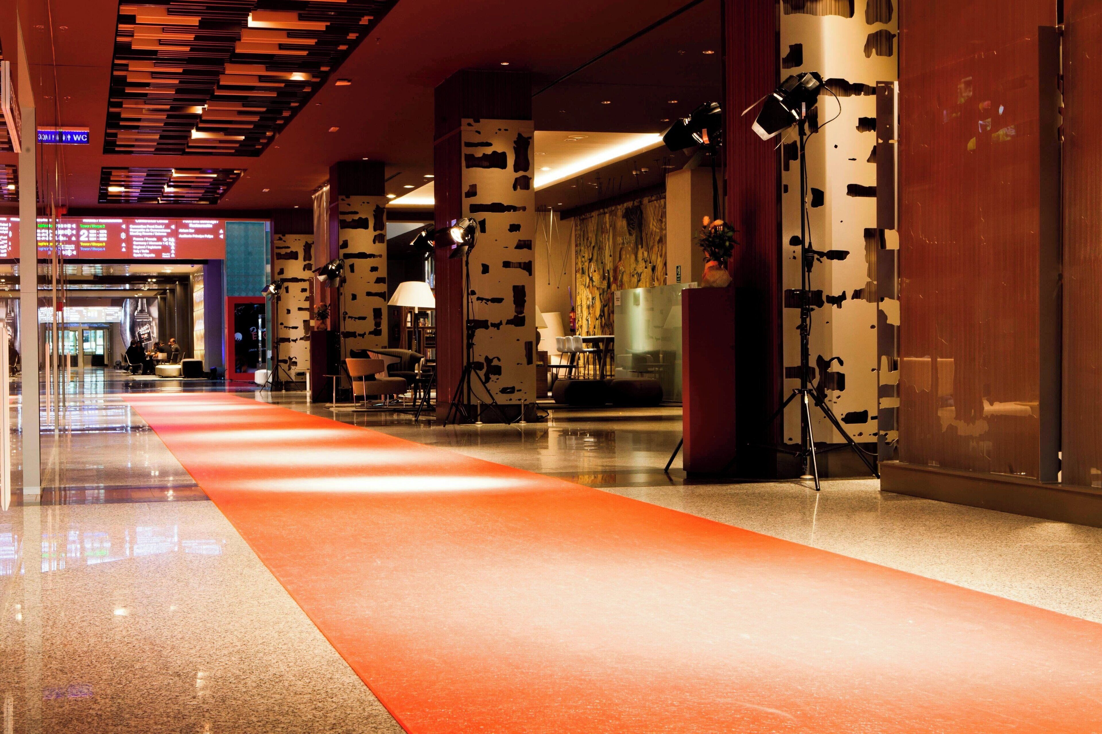 A modern hotel lobby with a long orange carpet, large columns, and a glass partition.