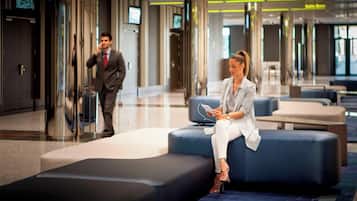 Un moderno vestíbulo de hotel con zona de estar, un ascensor de vidrio y una pantalla informativa digital.