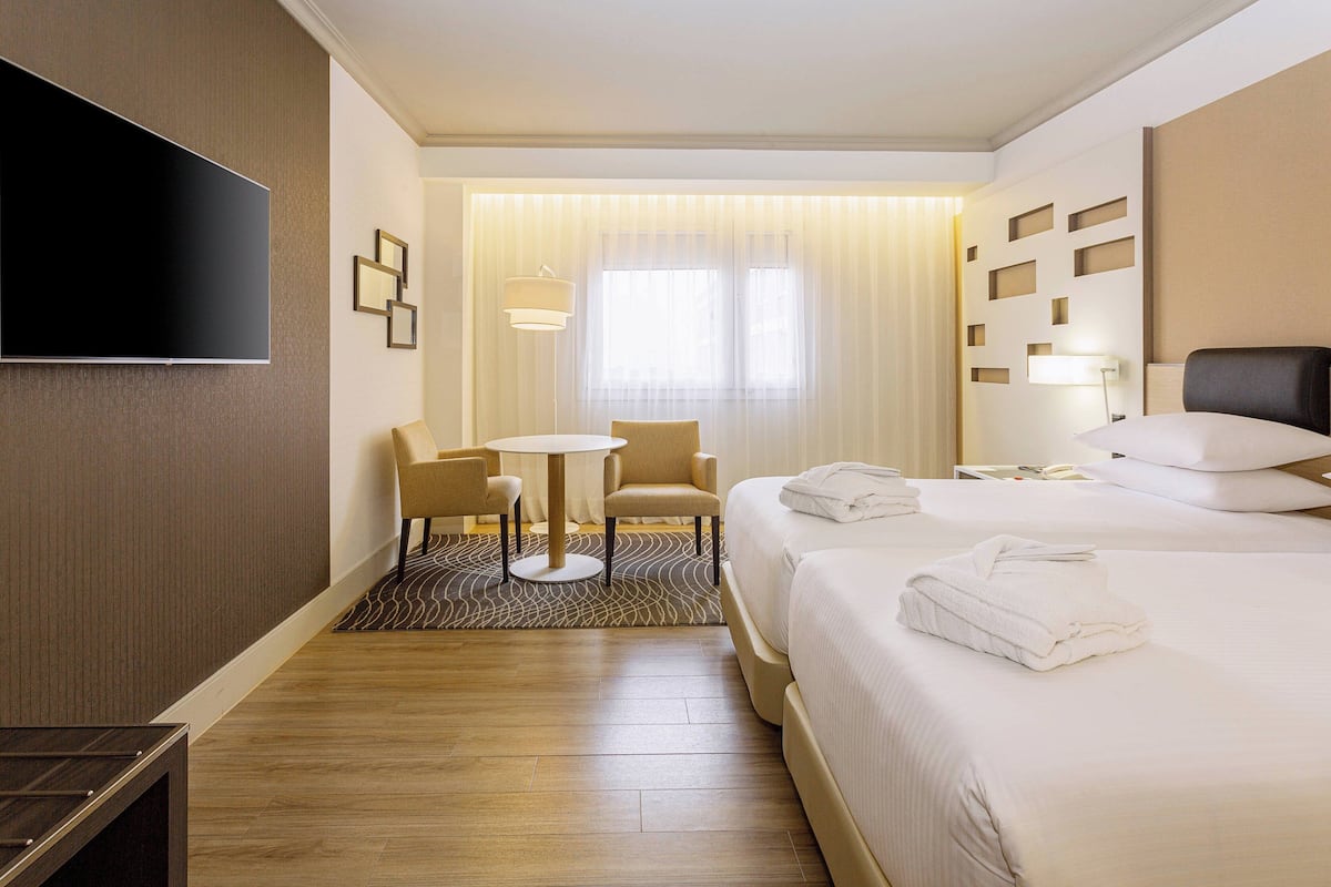 Ein Hotelzimmer mit Bett, Fernseher, Schreibtisch mit Stühlen, einem kleinen Tisch und einem Fenster mit Vorhängen.