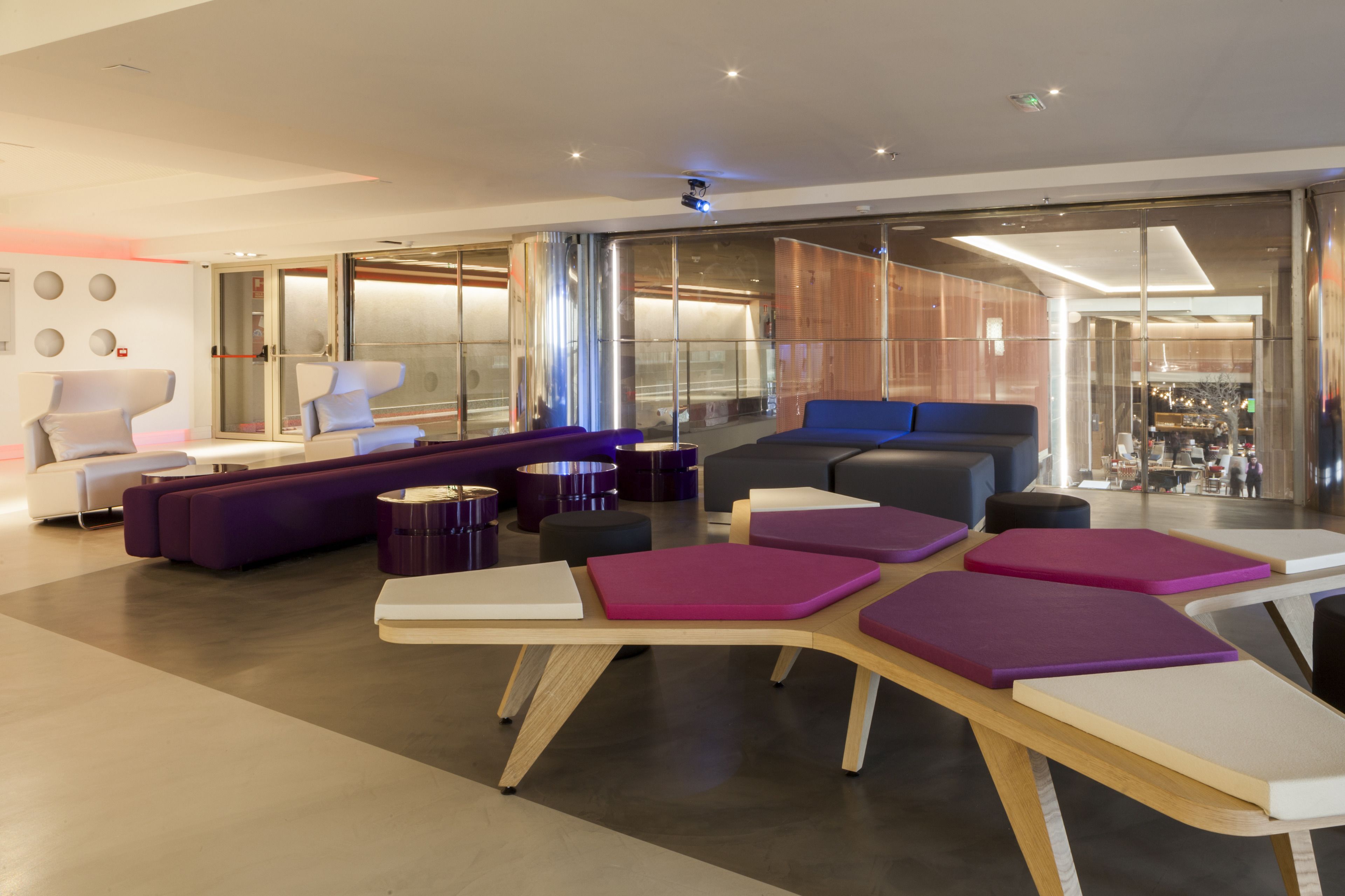 Een moderne lounge met zitplaatsen, waaronder banken en stoelen, een houten tafel met kussens en een glazen scheidingswand.