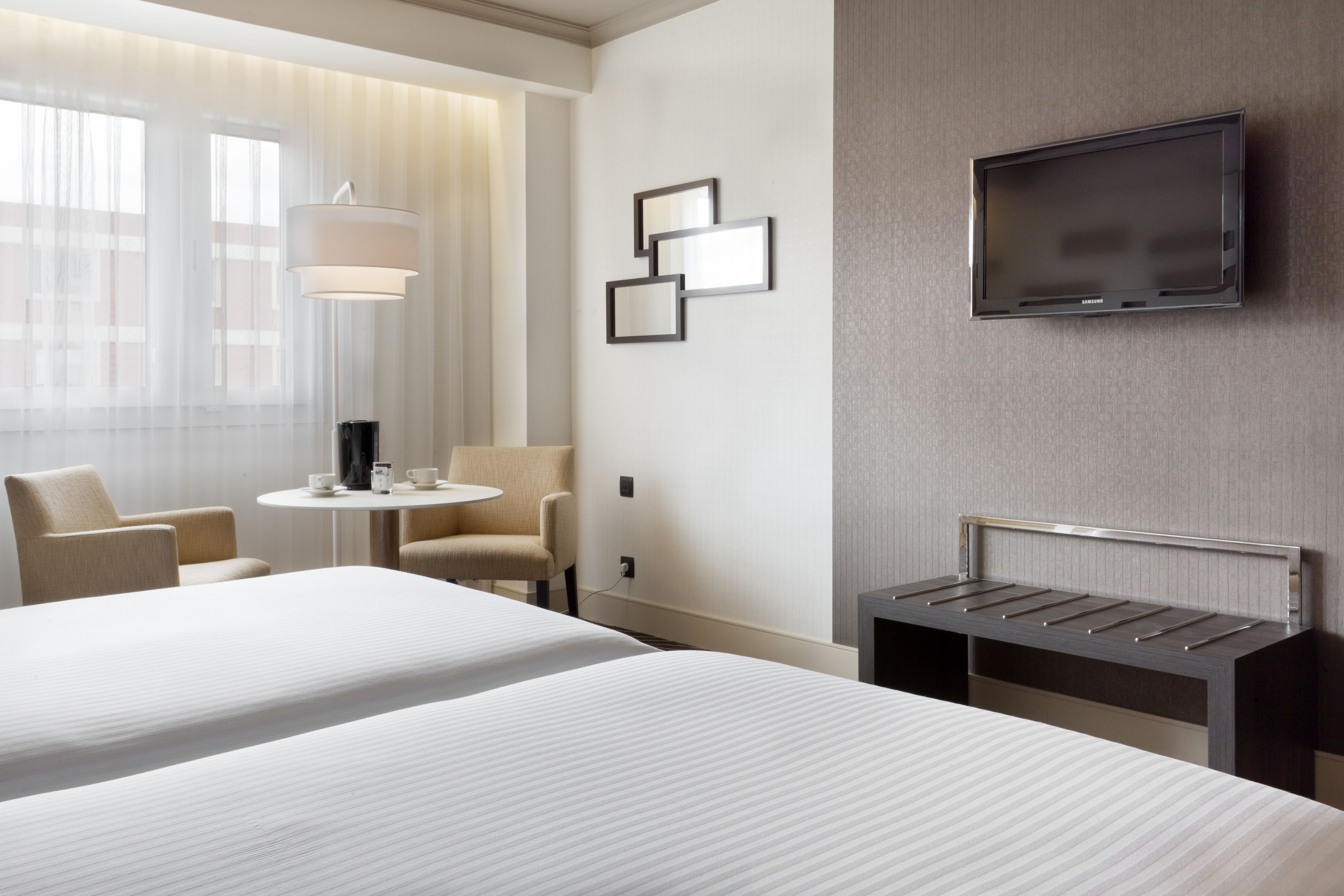 Habitació d'hotel amb dos llits, una televisió penjada a la paret, una taula petita amb una làmpada i una cadira.
