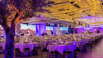 Un salón de banquetes con varias mesas redondas, cada una con mantel blanco y sillas, y un gran árbol en primer plano.