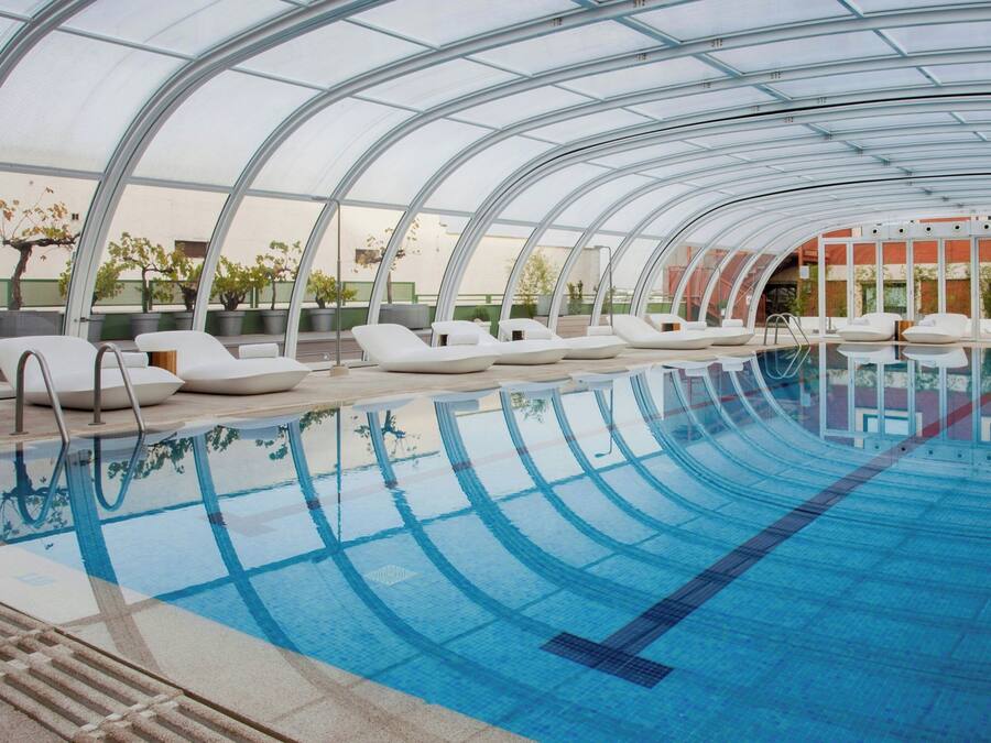 Una piscina interior moderna con techos de vidrio transparente y sillones blancos para relajarse.