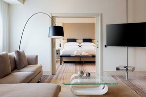 Ein modernes Hotelzimmer mit einer Couch, einem Glastisch, einem Flachbildfernseher, einem Bett mit Kissen und einem Nachttisch.