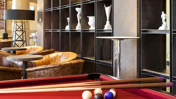 Una mesa de billar con bolas y tacos, un sofá y una lámpara en una habitación con paredes oscuras y estanterías llenas de trofeos.