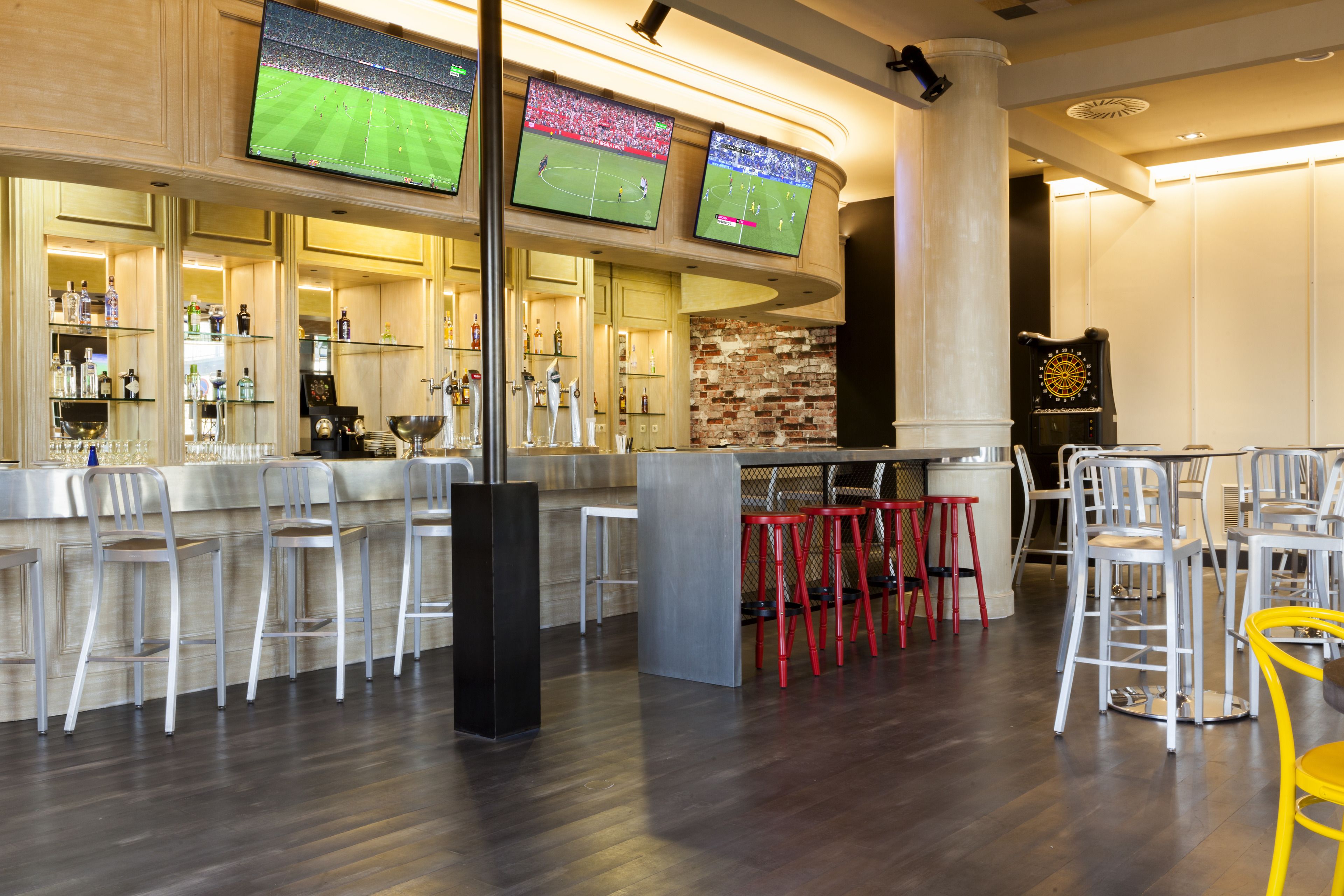 A bar area with multiple screens displaying sports events, bar stools, and a mix of red and yellow chairs.