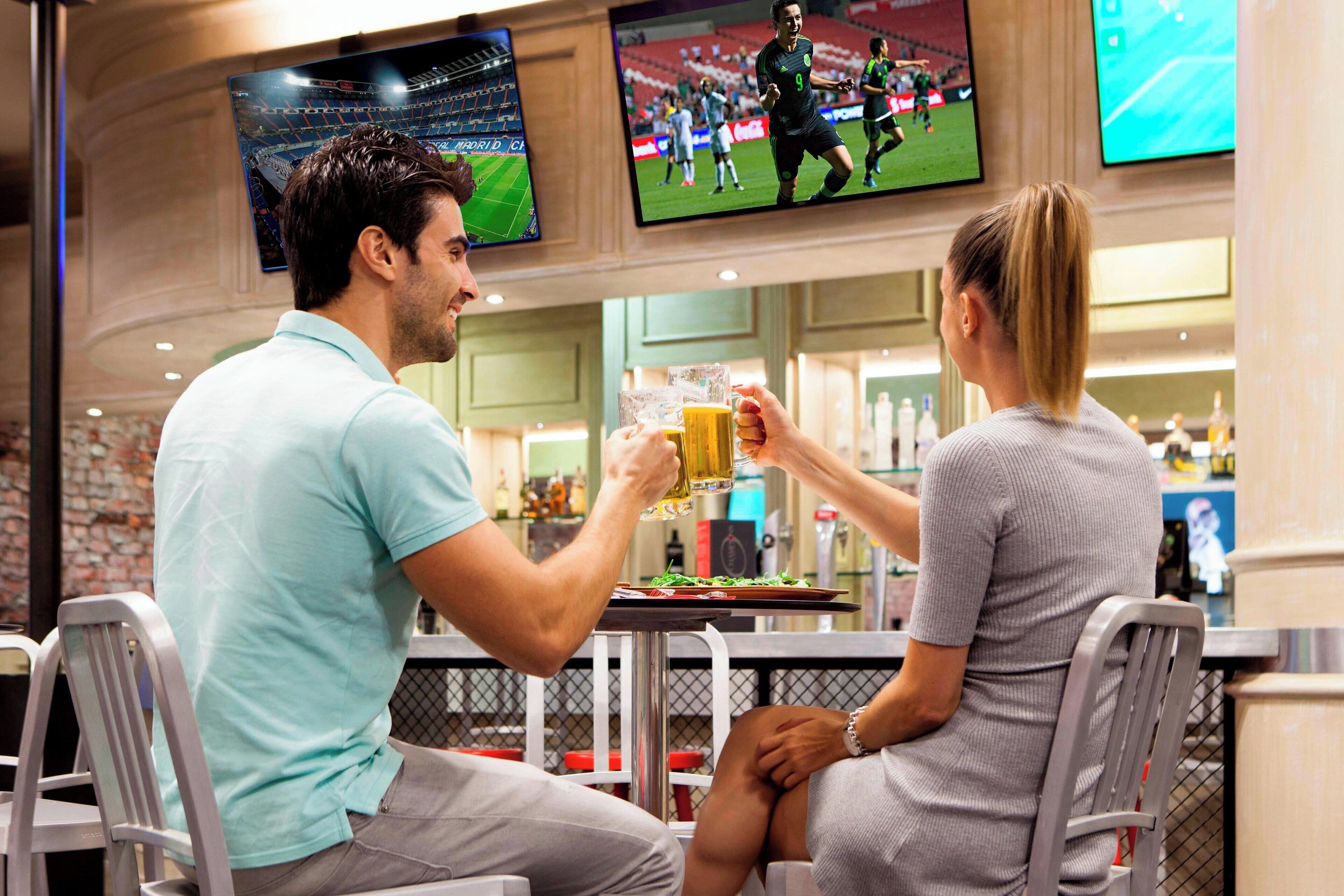 Un hombre y una mujer sentados en un bar con varias pantallas mostrando eventos deportivos.