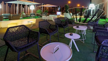 Un espacio al aire libre con mobiliario moderno, que incluye sillas y mesas, y una zona de bar con televisor de pantalla plana.
