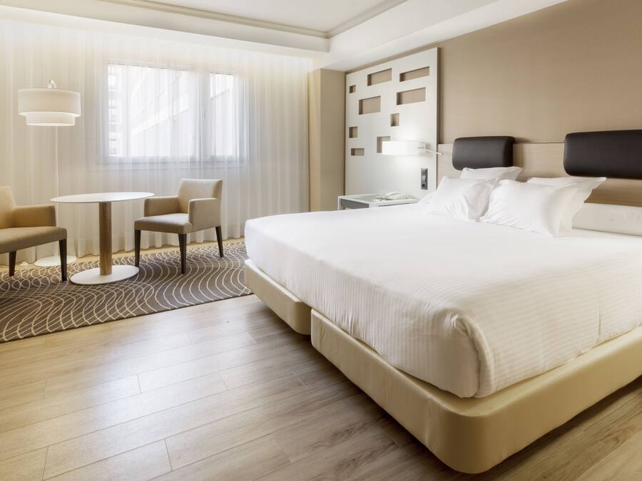 Een moderne hotelkamer met een groot bed, een klein rond tafeltje, twee stoelen, een bureau en een groot raam met dunne gordijnen.