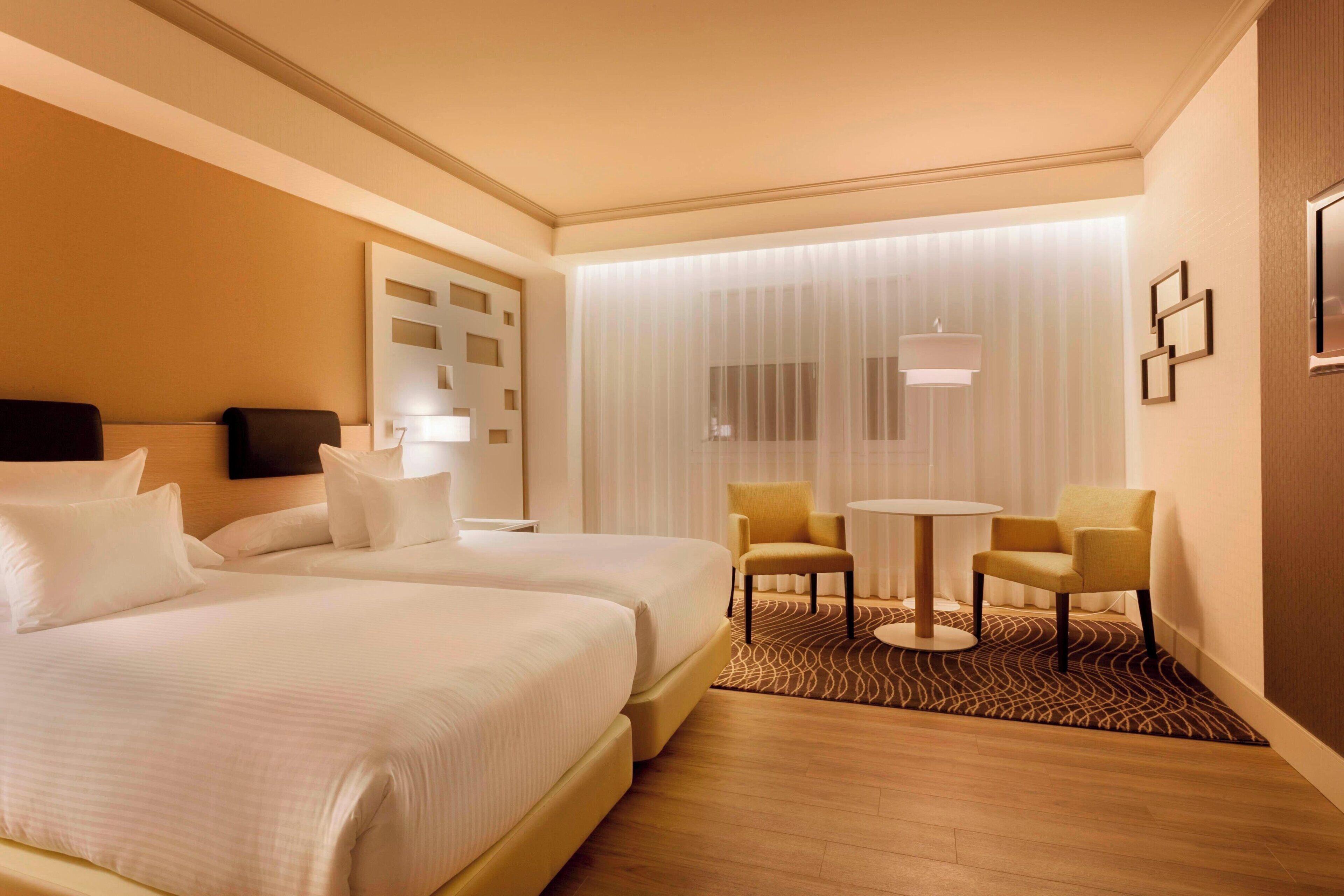 A hotel room with two beds, a small round table, two chairs, a lamp, and a large window with sheer curtains.