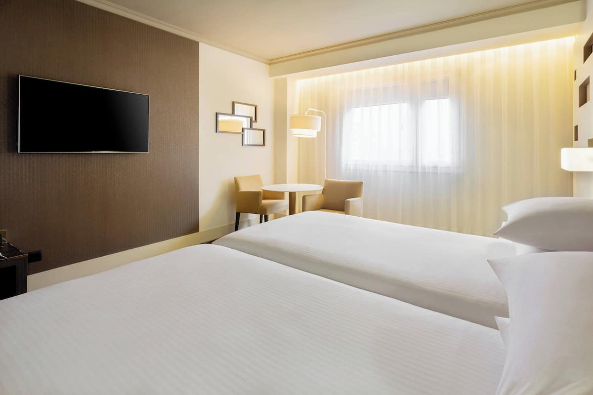 Ein Hotelzimmer mit zwei Betten, einem wandmontierten Flachbildfernseher, einem kleinen Tisch mit Stühlen und einem Fenster mit Vorhängen.
