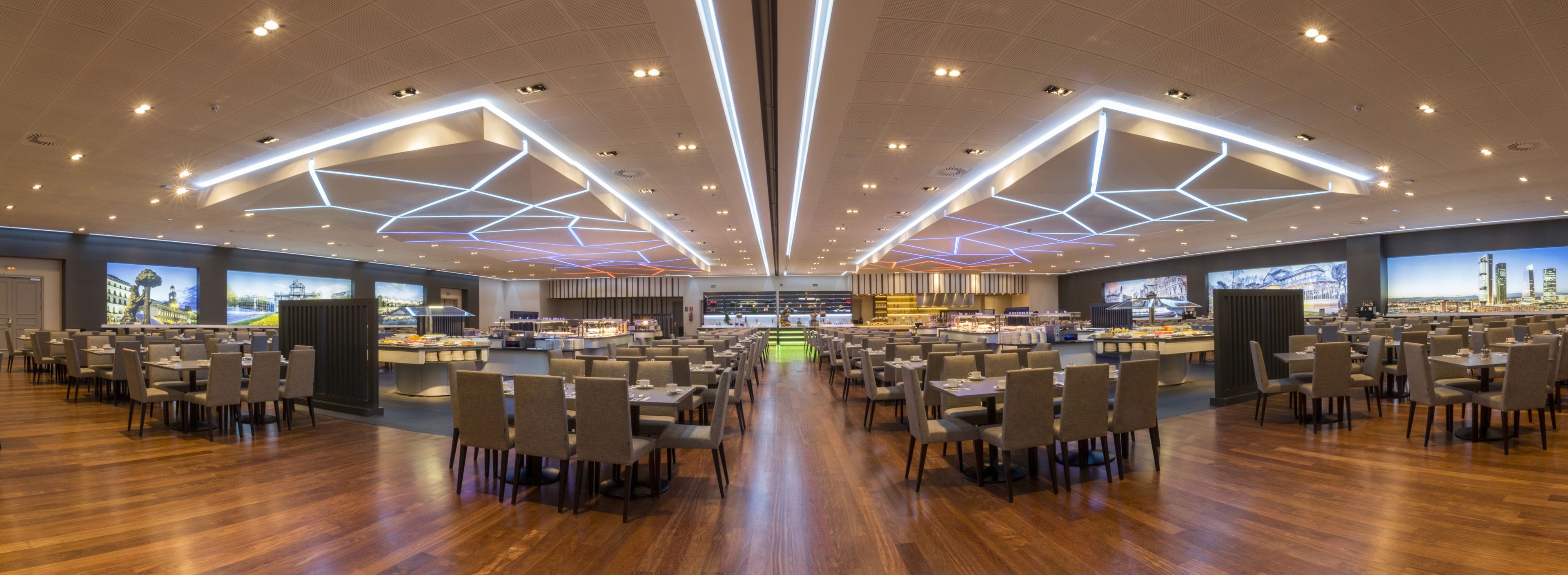 A spacious dining area with multiple tables and chairs, a central buffet, and large screens displaying cityscapes.