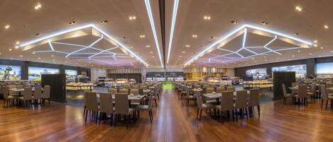 A spacious dining area with multiple tables and chairs, a central buffet, and large screens displaying cityscapes.