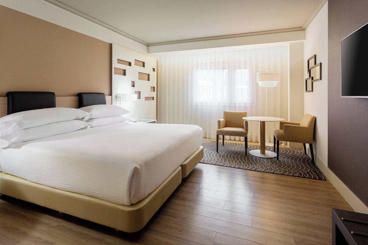 Ein Hotelzimmer mit einem großen Bett, einem Schreibtisch mit zwei Stühlen, einem Fernseher und einem Fenster mit Vorhängen.