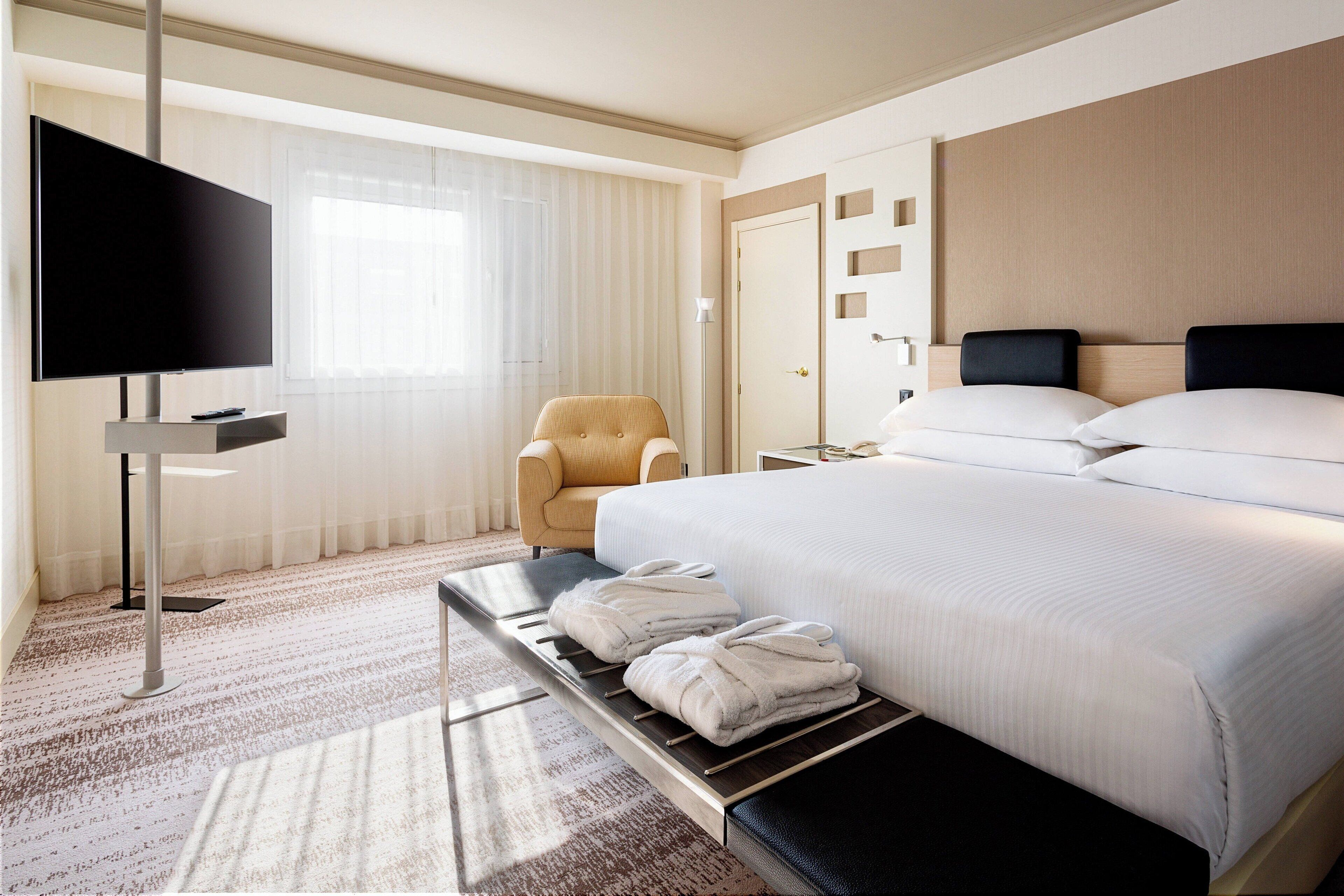Habitación de hotel con una cama grande, un televisor de pantalla plana, una silla y un banco con toallas.