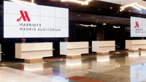 Moderni vastaanottoaula, jossa on suuri kyltti, jossa lukee Marriott Madrid Auditorium. Tilan sisustuksessa on käytetty minimalistista tyyliä, ja siinä on valkoiset vastaanottotiskit ja punaiset kukat.