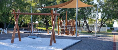 A playground with swings and a climbing structure, surrounded by trees and a covered seating area.