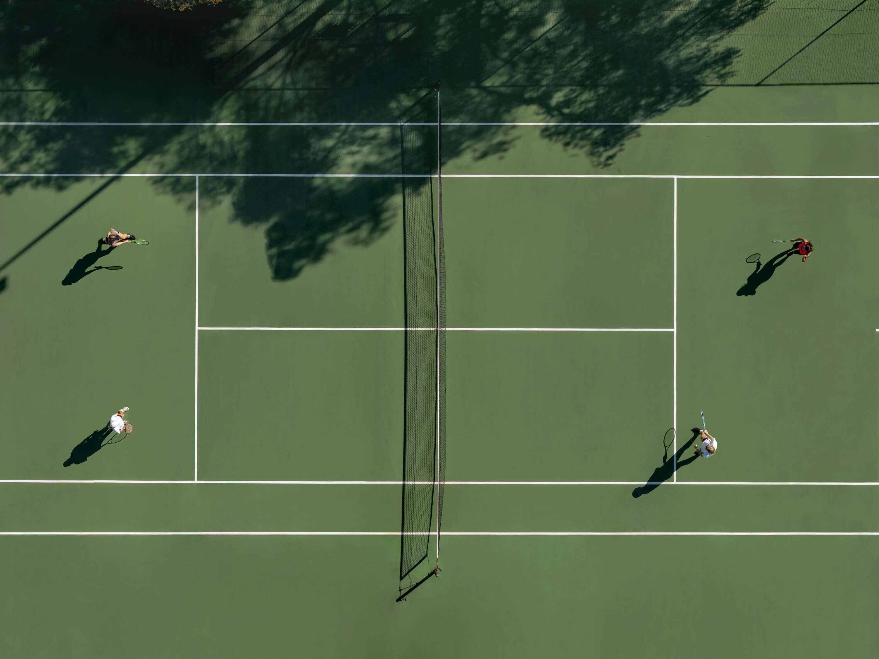Tenniskenttä, jossa neljä pelaajaa on pelaamassa, verkko ja taustalla puu.