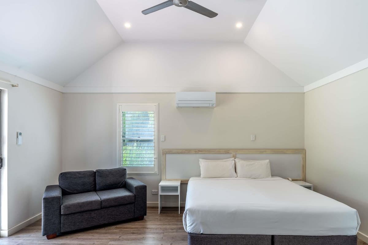 Un dormitorio con una cama grande, un sofá gris, un ventilador de techo y una ventana con persianas.