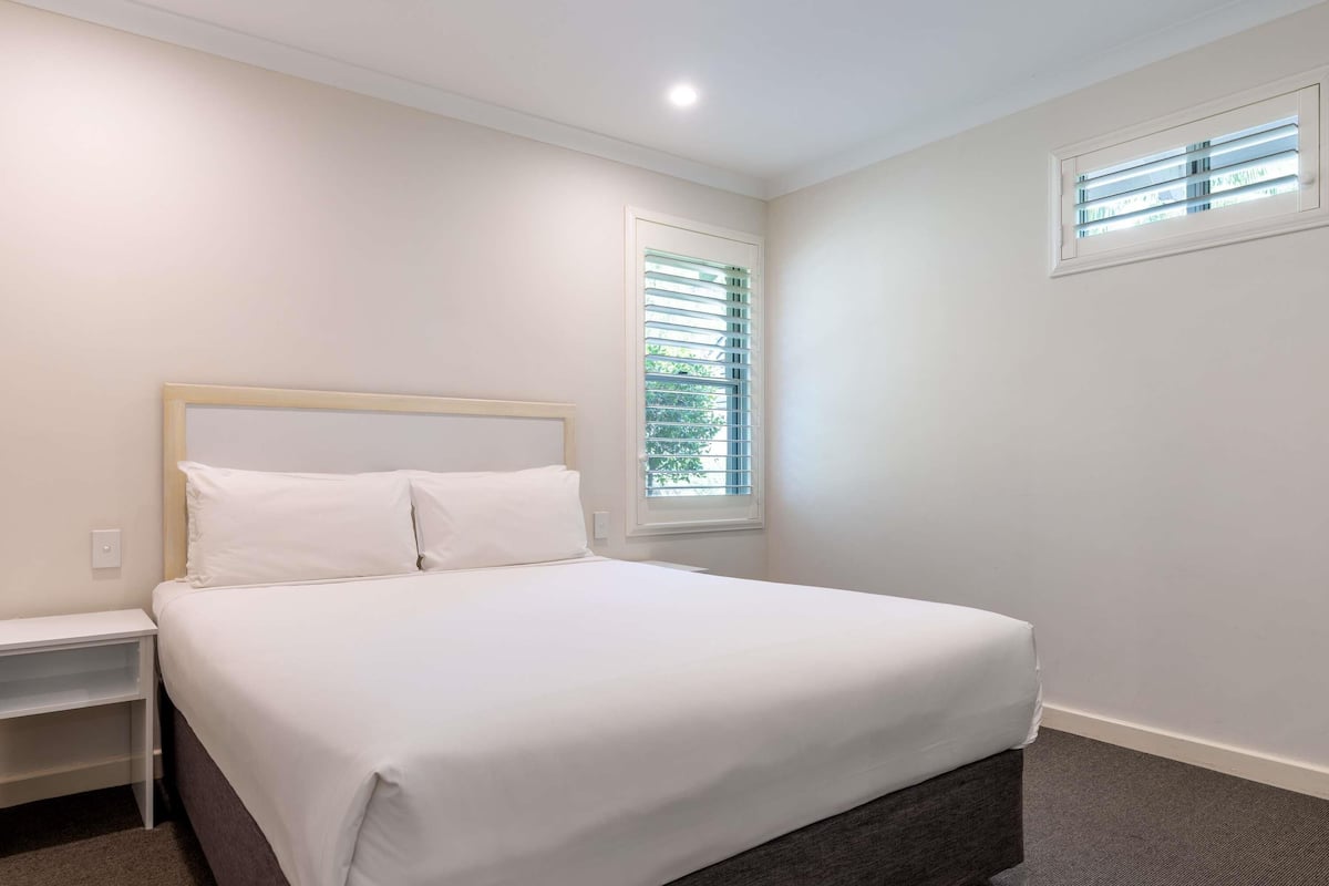 Un dormitorio ordenado con cama, mesita de noche, ventana con persianas y luz de techo.