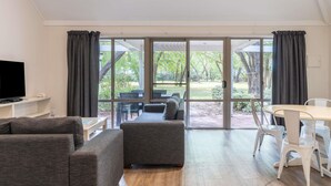 A modern living room with a flat-screen TV, grey sofas, a dining table with white chairs, and large sliding glass doors leading to an outdoor area. - Bayview Geographe Resort by Accor (Broadwater)