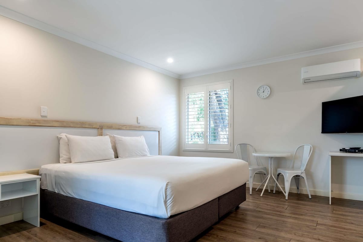 Habitación de hotel con cama, escritorio, silla, televisor y ventana con persianas.