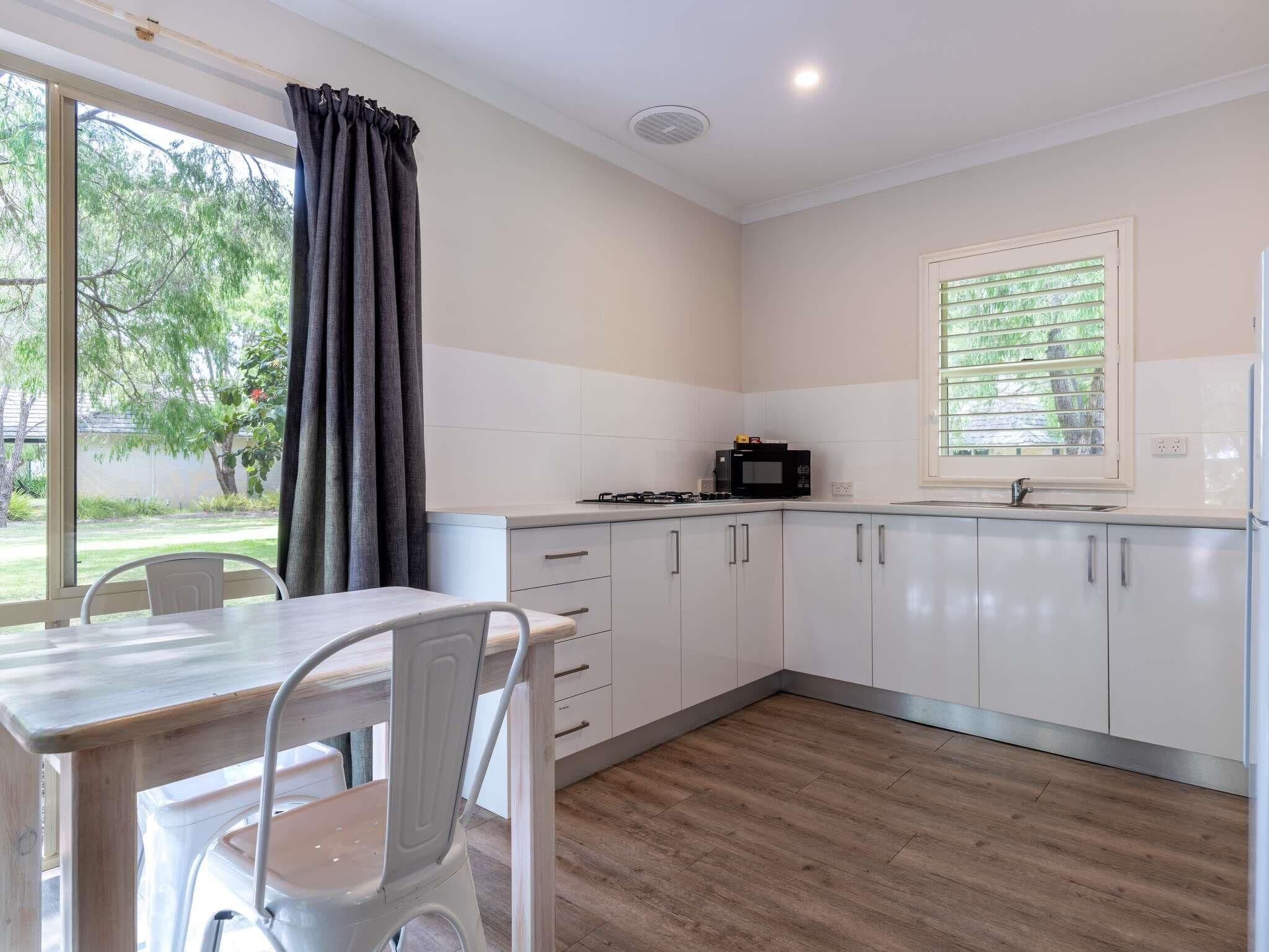 A modern kitchen with white cabinets, a dining table with chairs, a window with blinds, and a view of greenery outside.