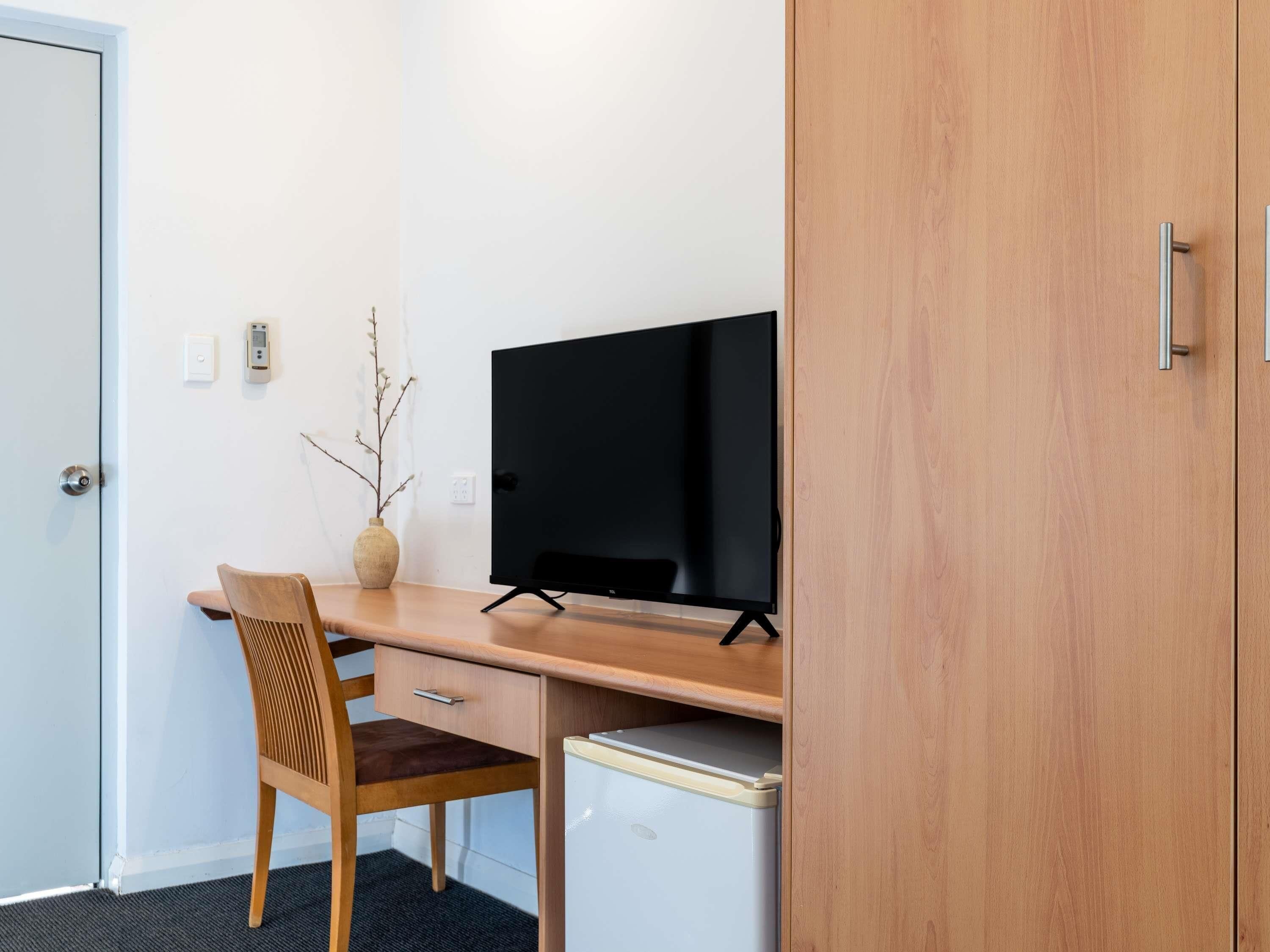 A compact room with a wooden desk, a television, a chair, a small refrigerator, and a door.