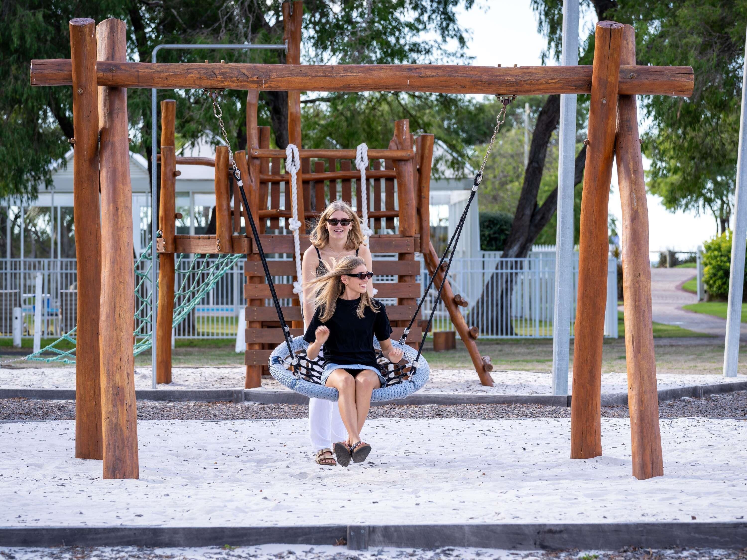 A wooden swing set with a metal frame, surrounded by a fenced area with trees and a pathway.