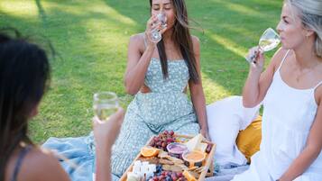 Trois personnes sont assises sur une couverture à l’extérieur, et un pique-nique est disposé devant elles, comprenant du fromage, des raisins et des boissons.