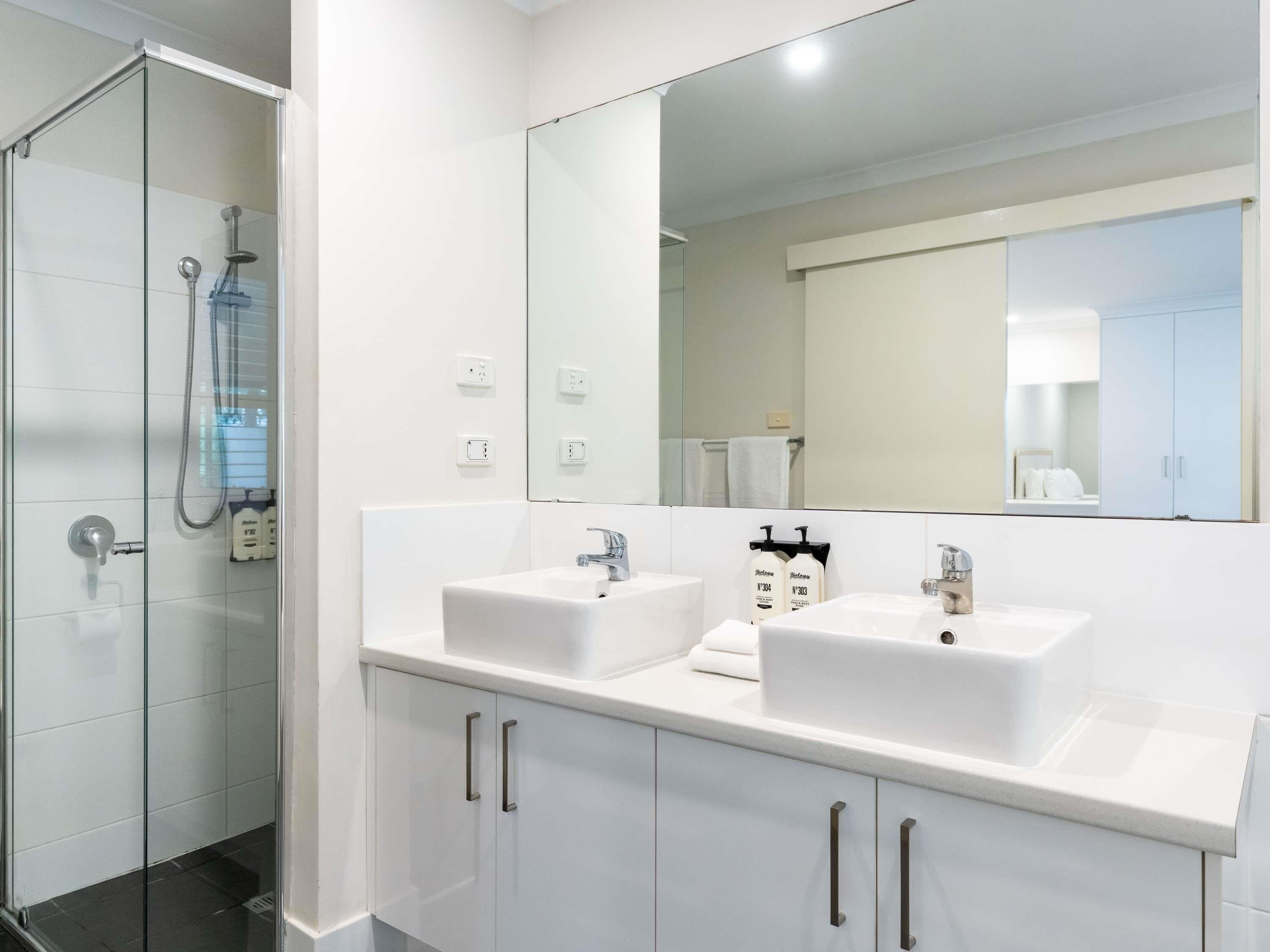Une salle de bain moderne avec une douche à parois de verre, deux lavabos, un grand miroir et des meubles blancs.