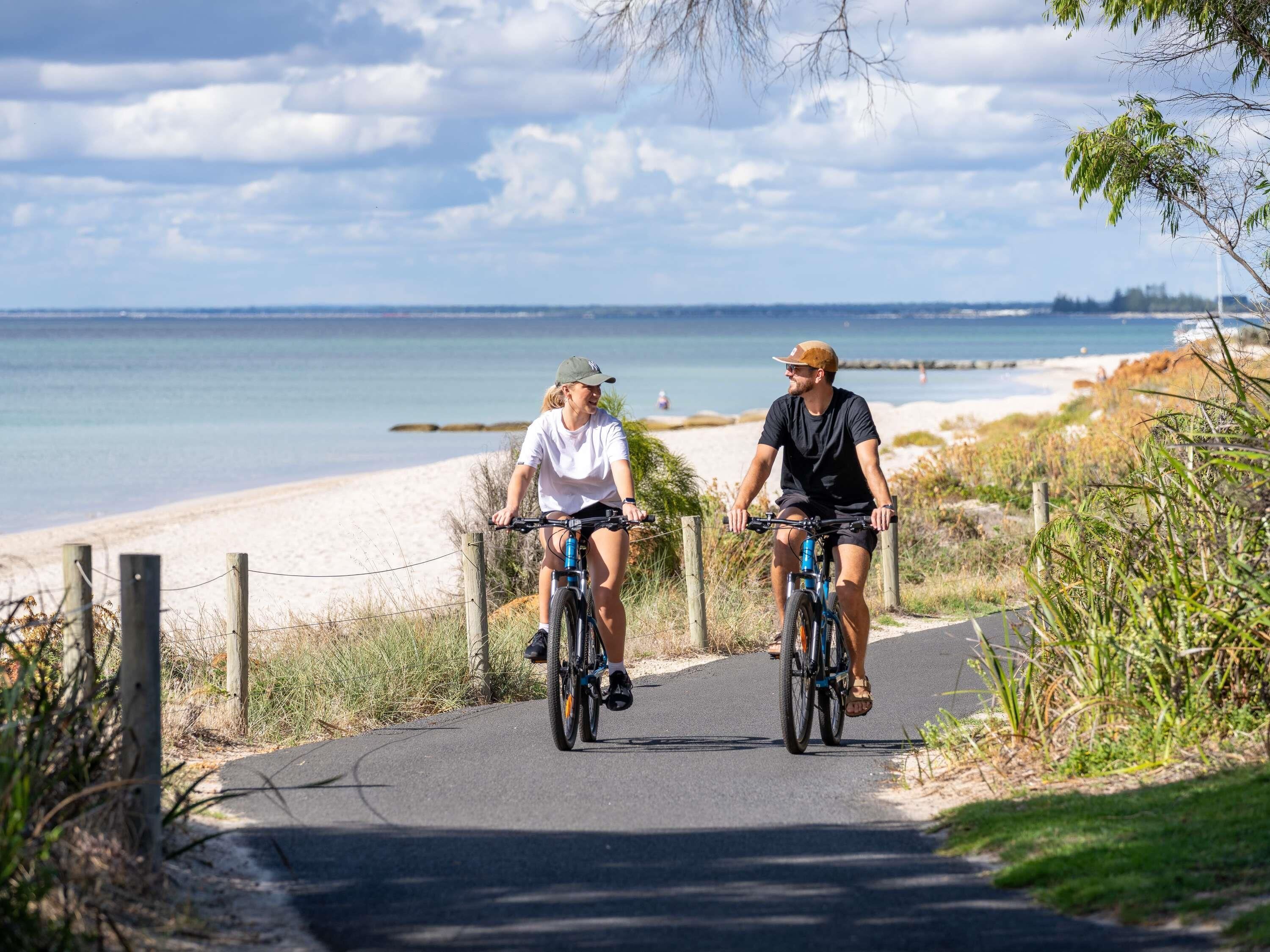 Two people riding bicycles on a paved path beside a beach with a view of the ocean.