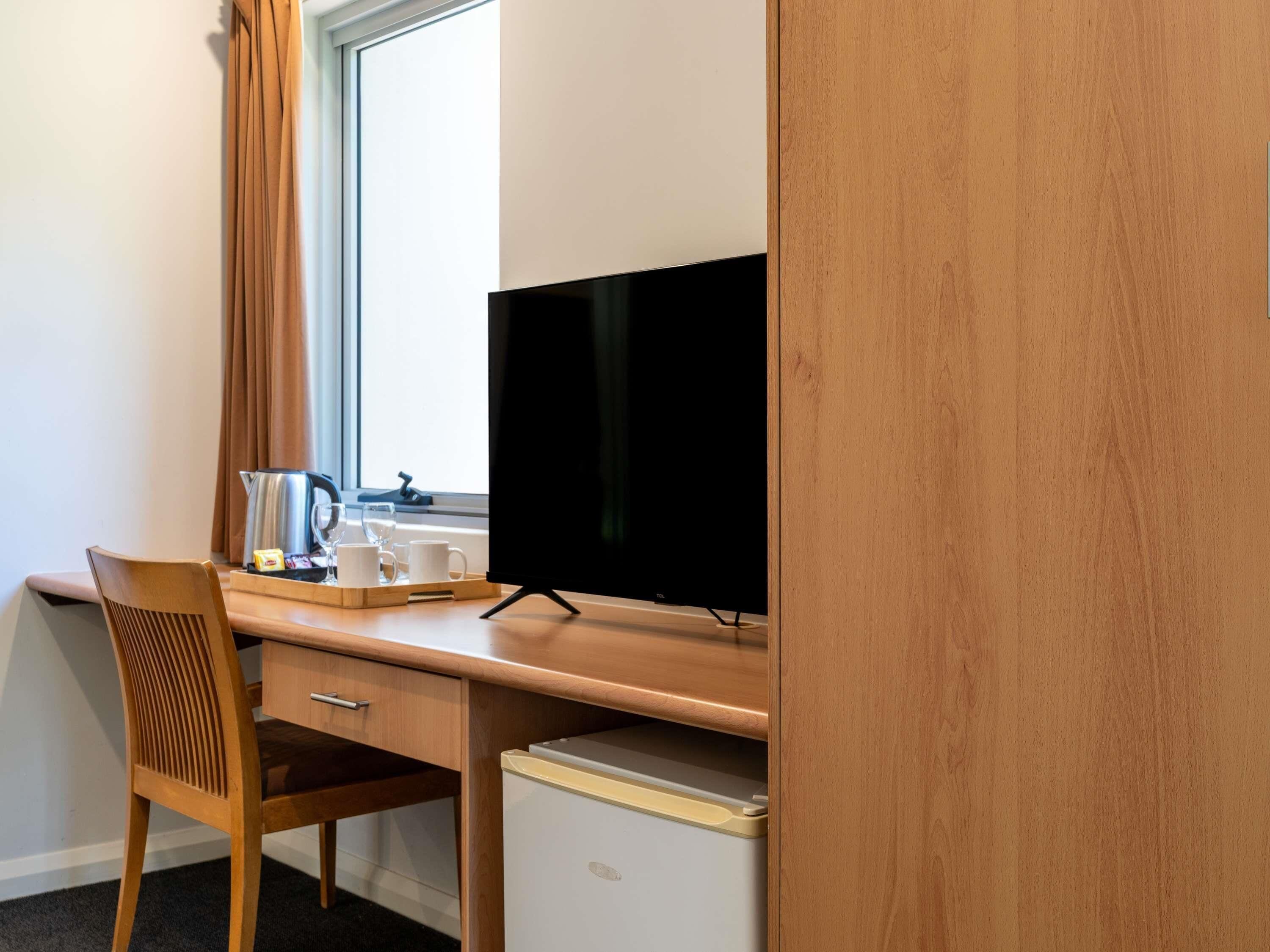 A hotel room with a wooden desk, a television, a mini-fridge, a chair, and a window with curtains.