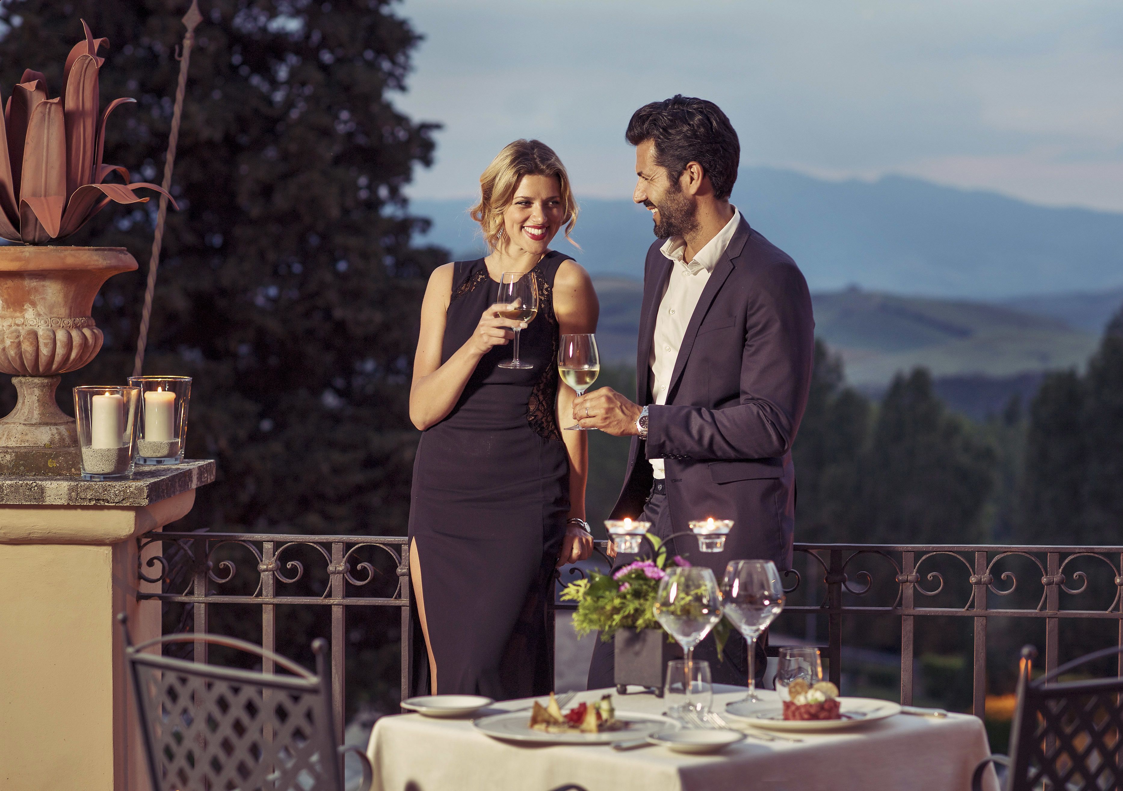 A couple dining at an outdoor table with a scenic view, candlelit table setting, and a decorative vase.