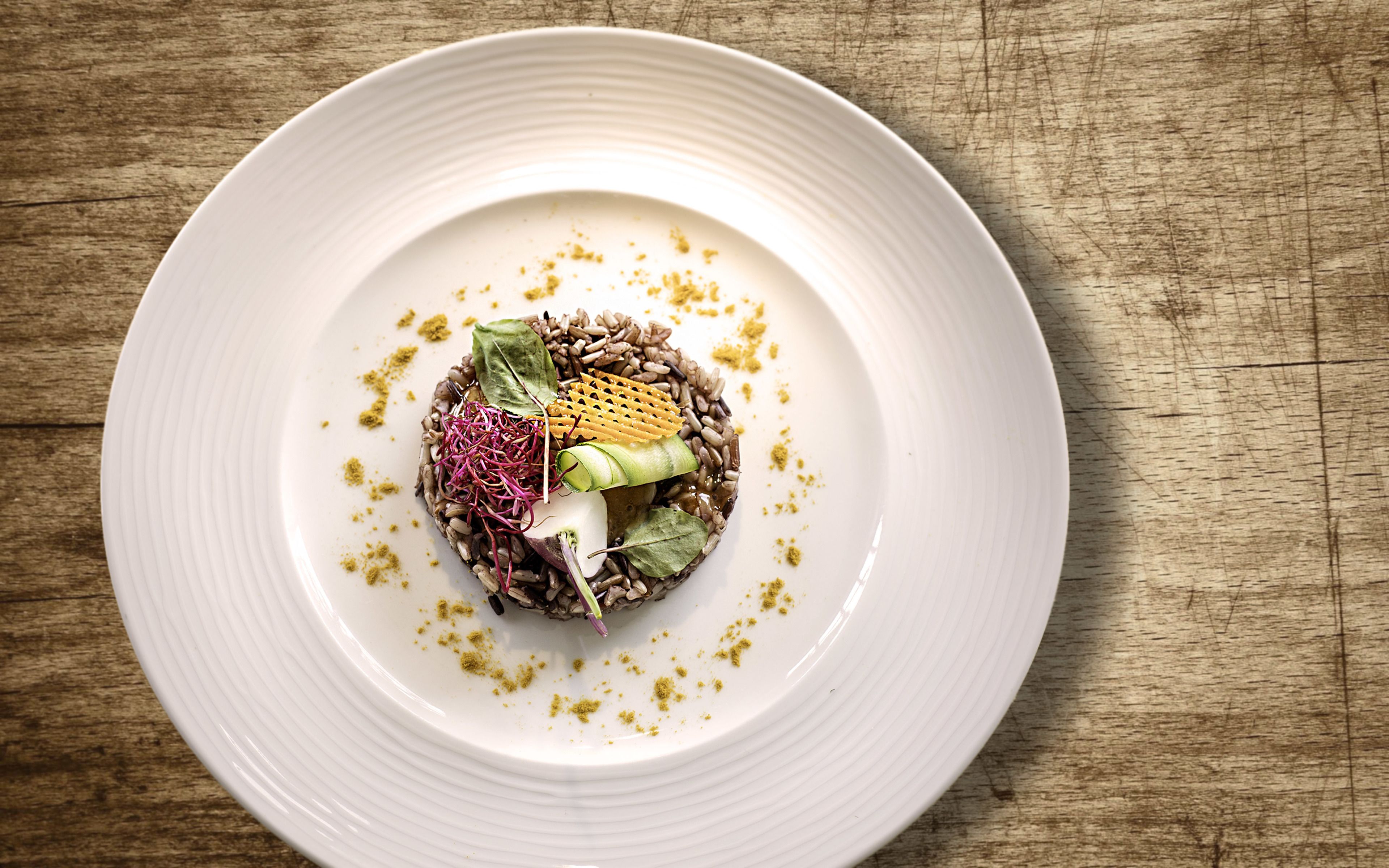 Une assiette blanche ornée d’une composition centrale de céréales, de graines et de légumes verts, le tout entouré de quelques céréales éparpillées et de quelques feuilles.
