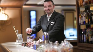 Un coin bar avec un barman en train de préparer un cocktail, un bar bien garni avec différentes bouteilles, et un téléviseur en arrière-plan.