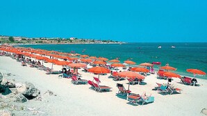 Une plage bordée de rangées de parasols orange et de chaises de plage, une mer d’un bleu limpide et des bâtiments en arrière-plan.